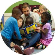 Preschool teacher sitting with 5 preschoolers around him reading a book.