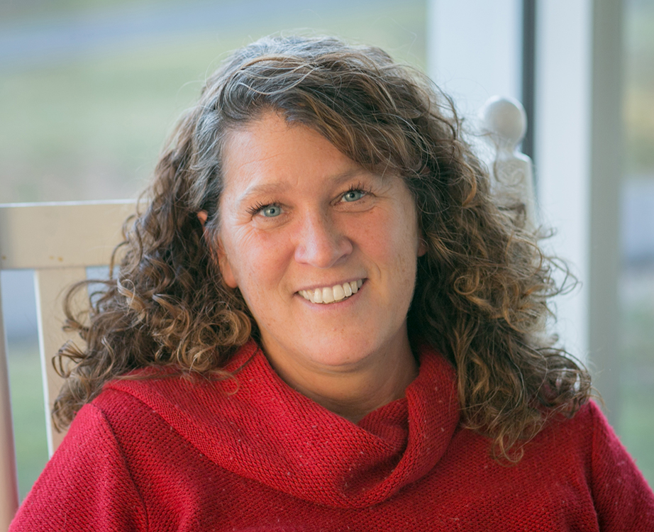 Jennifer Maeverde, a woman with long brown hair, blue eyes and a red knit sweater seated in a white wooden chair in an outdoor setting.