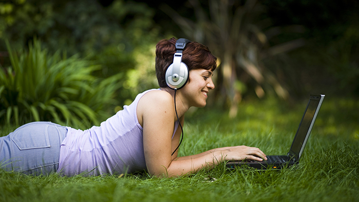 Young female student using laptop and headphones whilst lying on the grass.