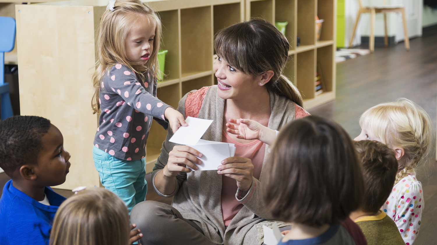 A preschool teacher sitting on the floor with a group of multi-ethnic children in a circle. They are watching her as she holds up a card. A little girl, a special needs child with down syndrome, is standing beside her handing a card to another child.