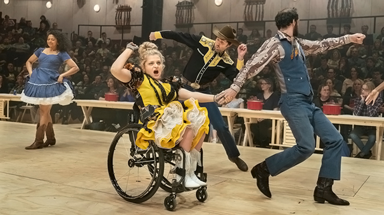 Actor Ali Stroker in a wheelchair pumping her fist with other actors dancing around her during Broadway revival of Oklahoma!