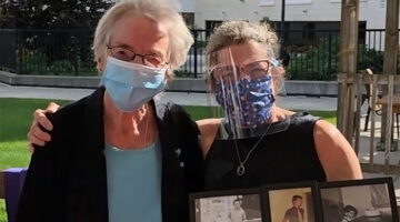 Two masked older women standing side-by-side outside. The woman on the right has her right hand on the other woman's shoulder and is holding a collage of framed family photos in her left hand.
