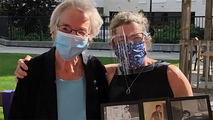 Two masked older women standing side-by-side outside. The woman on the right has her right hand on the other woman's shoulder and is holding a collage of framed family photos in her left hand.