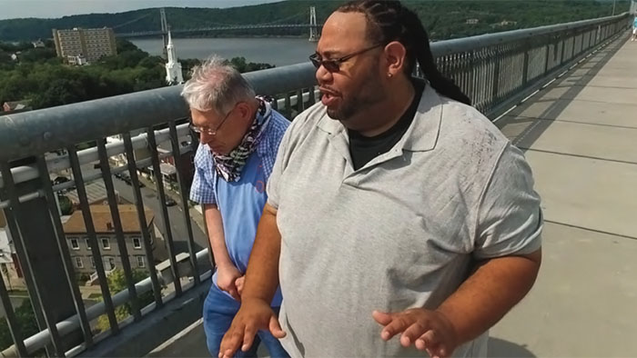 An African-American man and a senior white man with developmental disabilities walking along a pedestrian walkway overlooking a river.