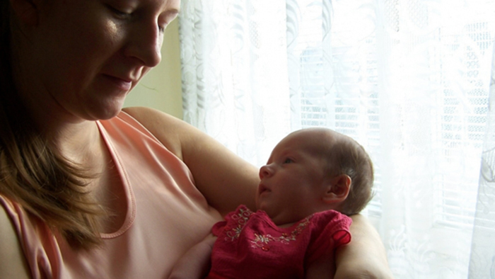 A young mother with long brown hair and a peach-colored top cradles her infant daughter in her left arm.