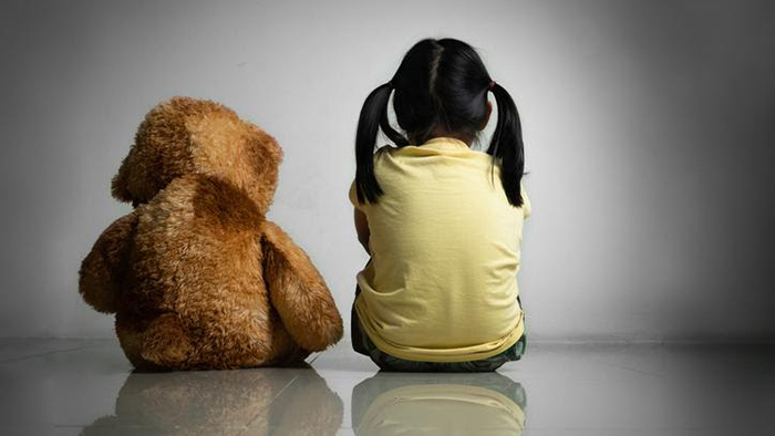 A stuffed brown bear and a young girl in a yellow shirt with black hair in pigtails sit with their backs to the camera.