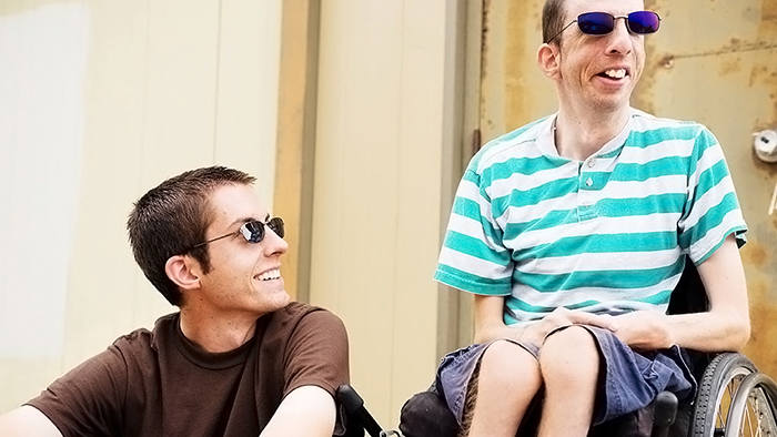 Adult twinbrothers istting next to each other: both are wearing sunglasses and smiling. One brother is seted on a step looking up at his twin, who has a developmental disability and uses a wheelchair.
