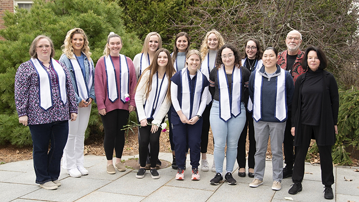 Thirteen people standing outside for a group photo. Two are college professors and the remaining 11 are college students wearing blue and white graduation stoles that signify their completion of a Disability Studies Minor.