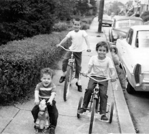 A black and white photograph from the 1960's showing three children who are siblings on a city sidewalk; a boy on a bicycle, a younger girl on a tricycle, and a younger boy on a self-propelled rocking horse with wheels.
