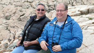 A smiling middle aged woman with short graying hair, wearing glasses, a black vest and blue jeans, sits on large rocks with her brother, a smiling middle-aged man with thinning hair, who is wearing glasses, a blue rain jacket and khaki pants with a pair of binoculars hanging from his neck.