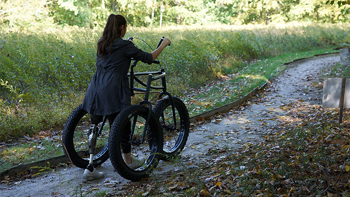 Woman with a prosthetic left leg exercising outdoors using an Afari 3-wheeled mobility device on a walking trail.