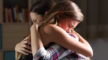 Side view of a grieving mother and teenage daughter embracing inside a home.